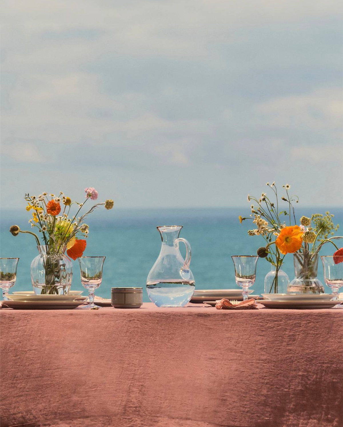 A dining table set against a beautiful turquiose ocean with clouds in the sky. The tablecloth is terracotta and there are beautiful plates, pitchers, glasses, and vases frilled with ranuncula flowers. 